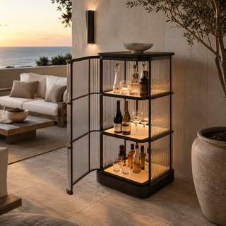 A three-tier black metal bar cabinet with ribbed glass rods and warm interior lighting, styled with bottles and glasses in an outdoor patio setting at sunset.