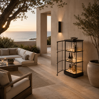 A black metal and glass three-tier glass rods cabinet with warm interior lighting stands on a sunlit patio beside a beige lounge sofa, with the ocean and evening sky in the background.
