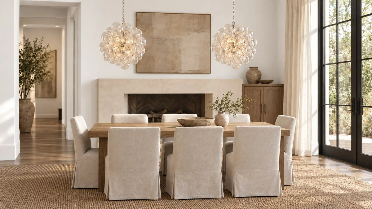 Two Spiral Bubble glass chandeliers over an oak dining table with linen slipcovered chairs and a plaster fireplace — organic modern dining room lighting