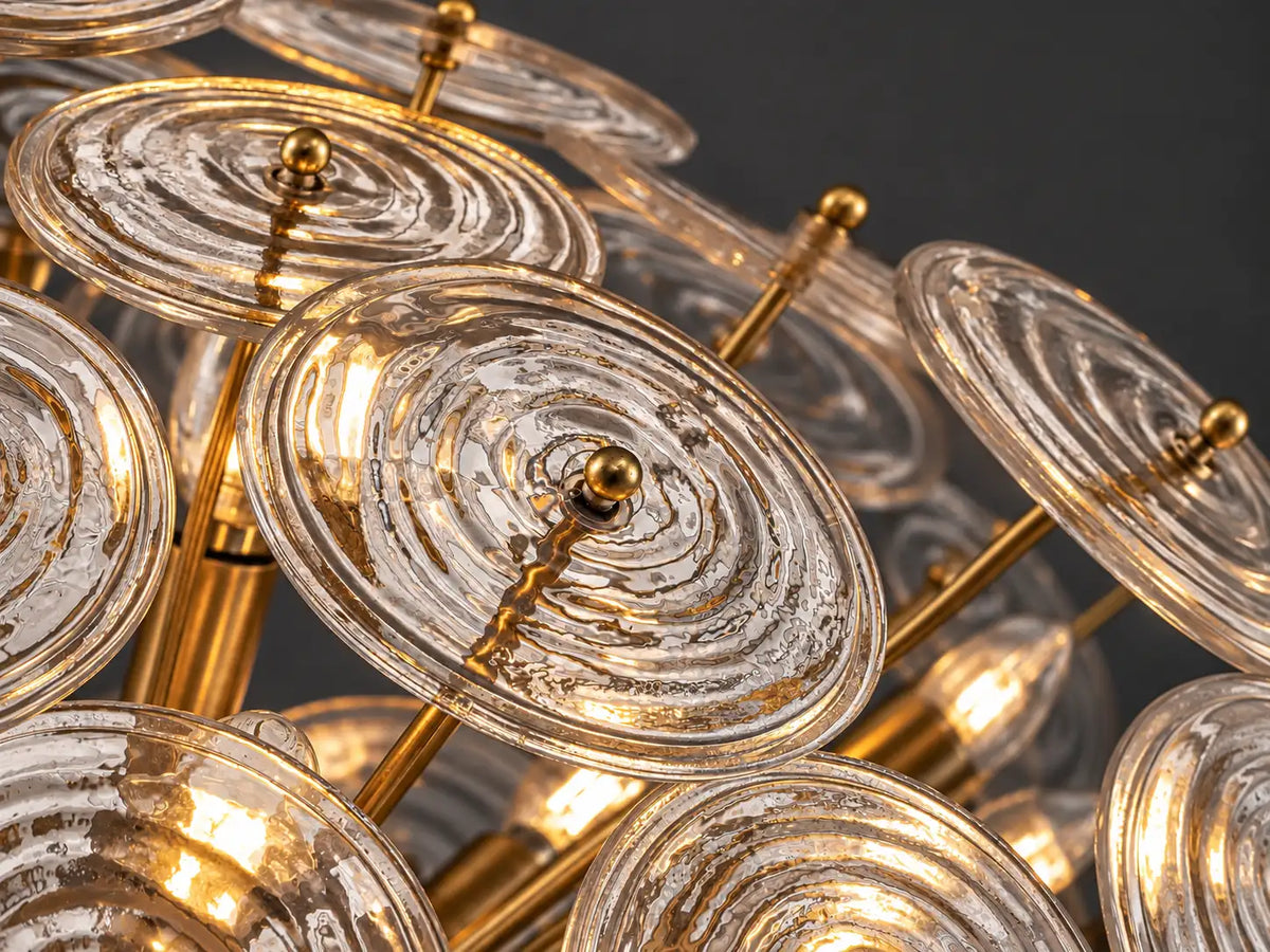 Close-up of a glass disc chandelier with clear, ripple-textured glass refracting warm light around brass-finish metal arms.