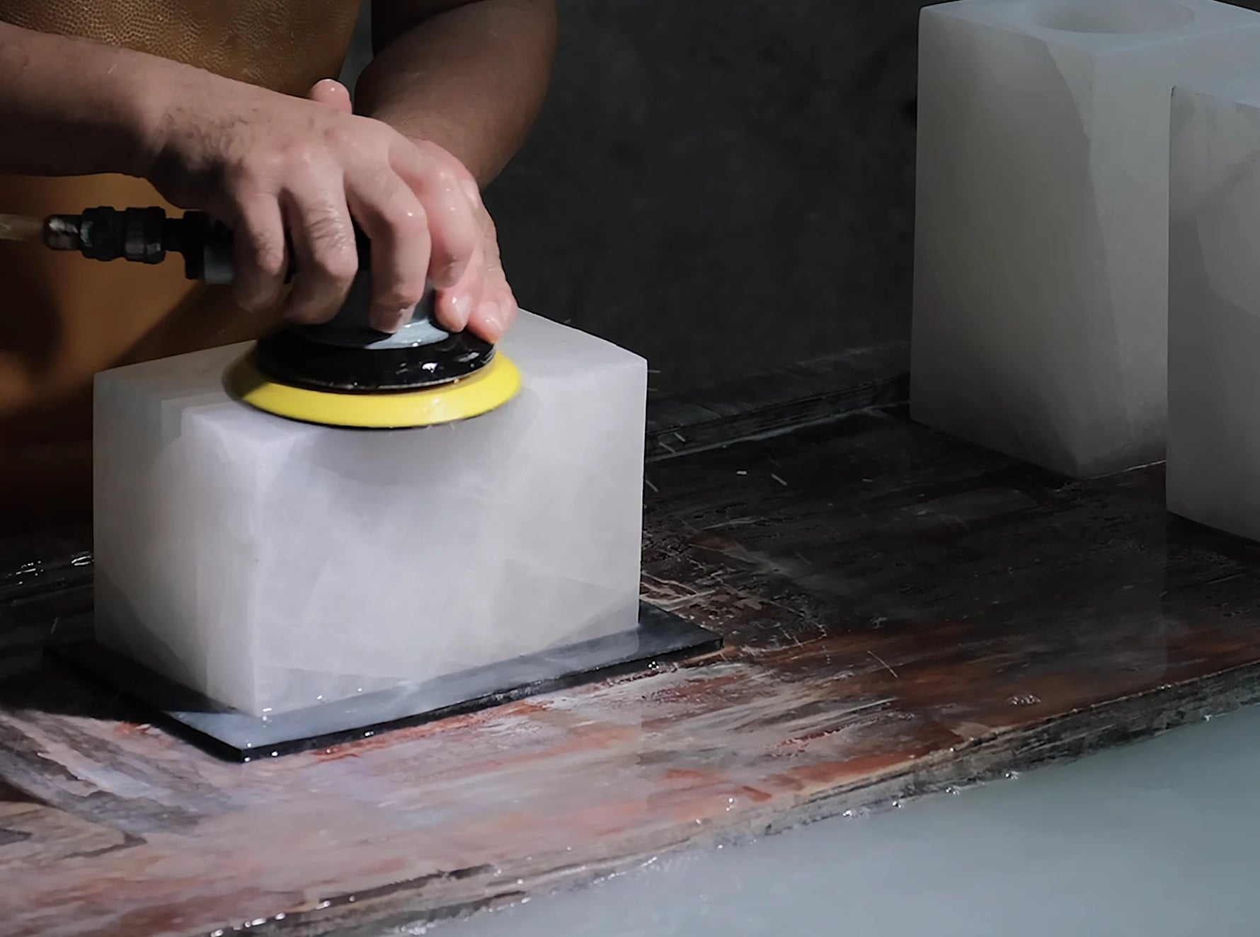 A person using a yellow and black rotary tool to polish a white rectangular calcite stone on a workbench, with more white calcite blocks in the background.