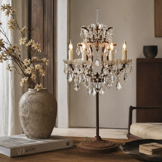 A vintage-style crystal chandelier table lamp with multiple candle-style bulbs and hanging clear glass teardrops, displayed on a wooden table in a softly lit interior setting next to a vase of flowers.