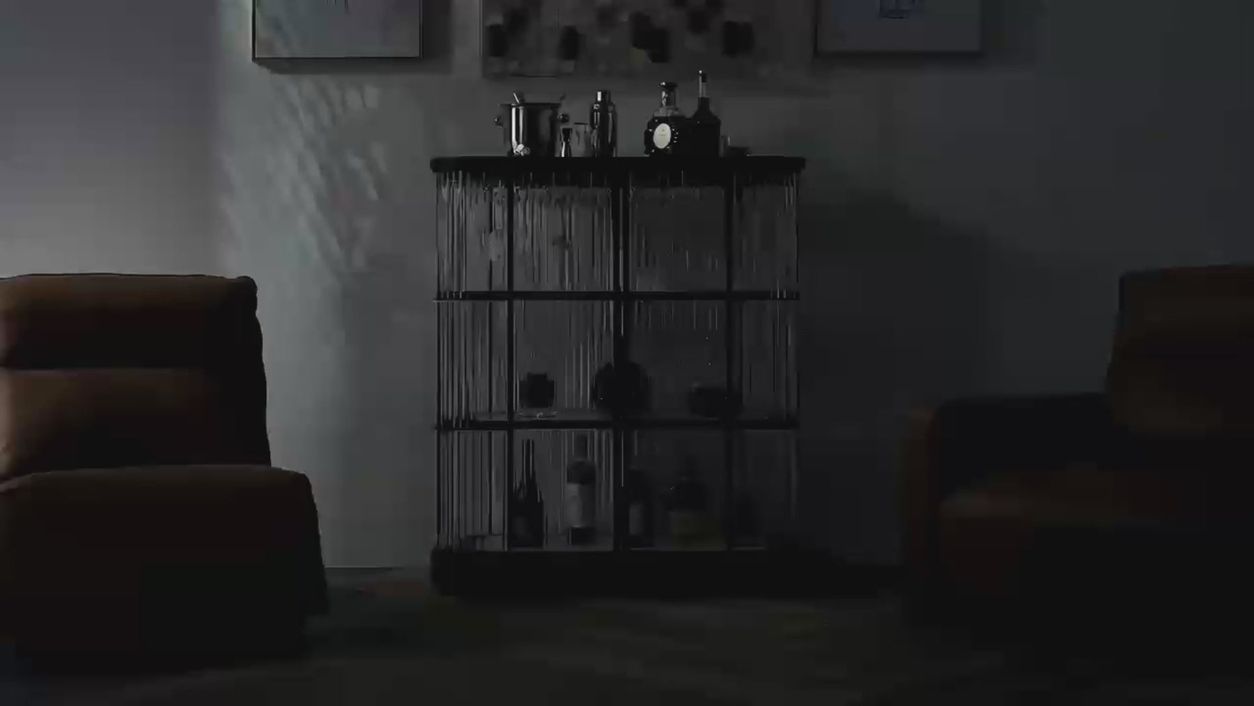 Moorizon Gatsby LED bar cabinet set between two leather chairs. The lighted liquor display cabinet has a black iron frame and clear glass rods, with glowing shelves holding bottles, crystal glasses and a stemware rack as a drink is poured.